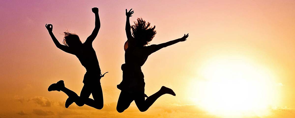 Zwei junge Menschen springen vor Freude und Glück in die Luft. Rechts ist die Abendsonne zu sehen.