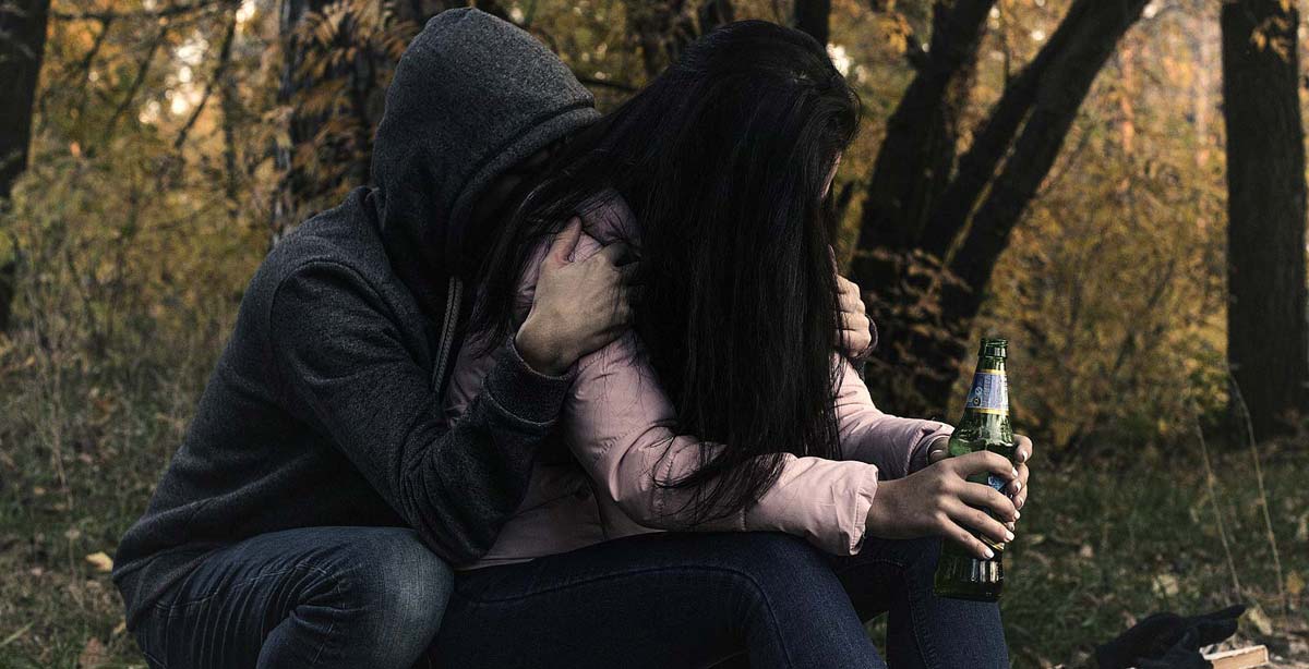 Alkoholsüchtiger Mann und Frau sitzen im Wald. Er versteckt sein Gesicht, sie hält eine Bierflasche in der Hand.
